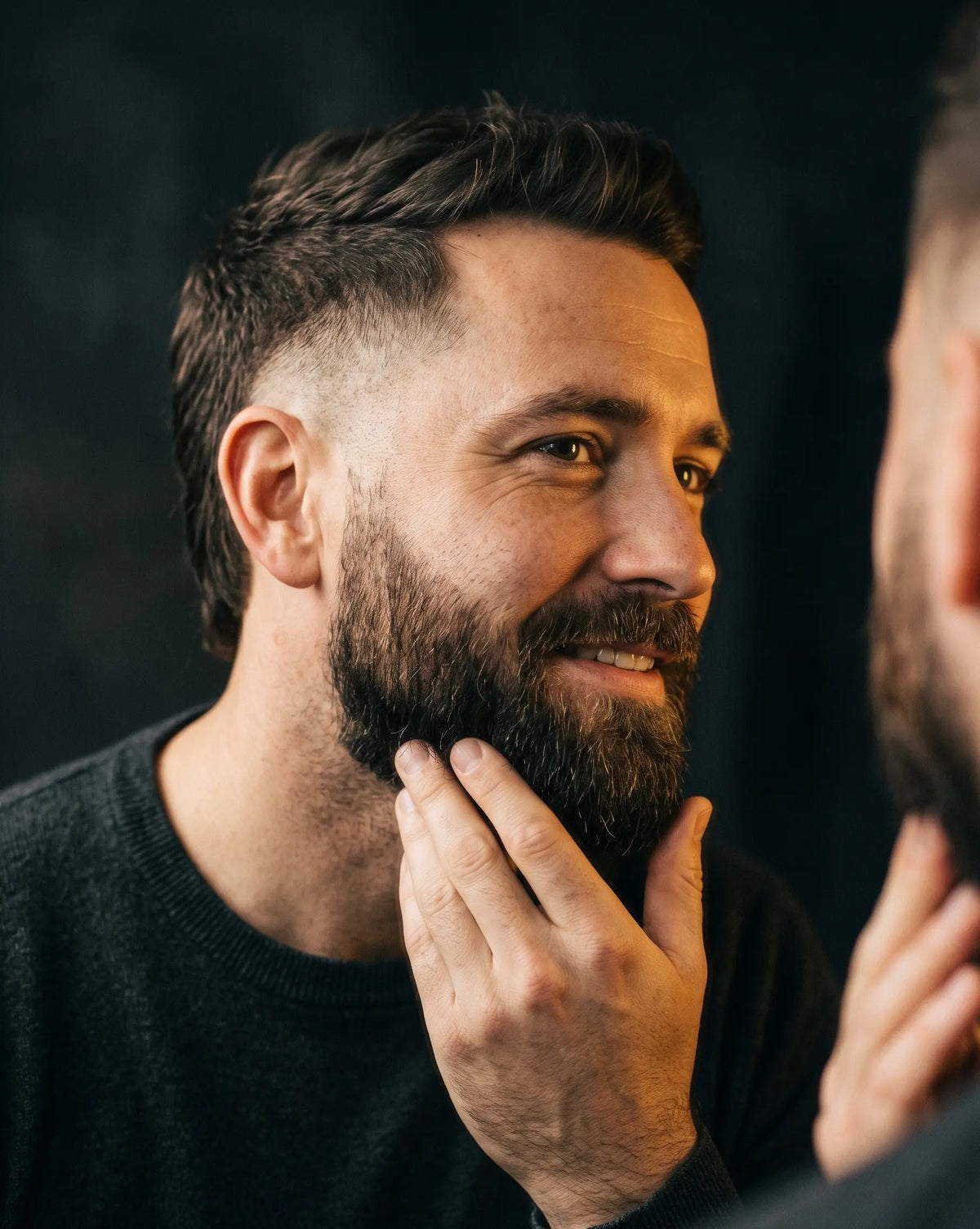 A close-up lifestyle shot of a man with a well-groomed dark beard touching his chin, looking into a mirror. The background is a dark, textured studio backdrop.