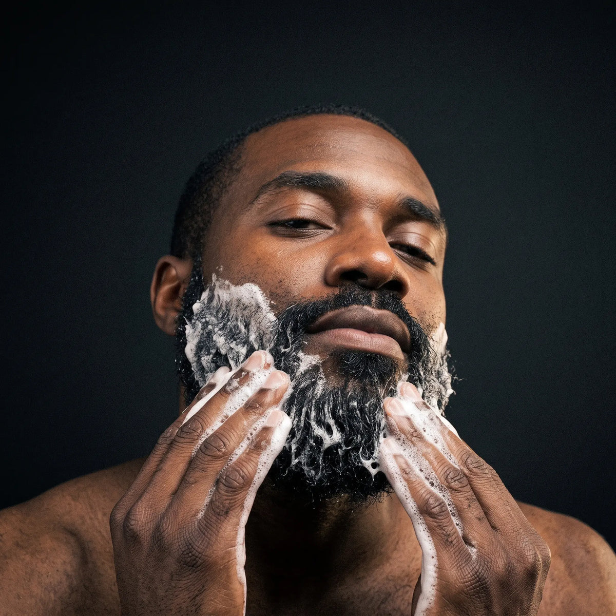 A close-up of a man with a beard washing it with white shaving foam against a dark background.