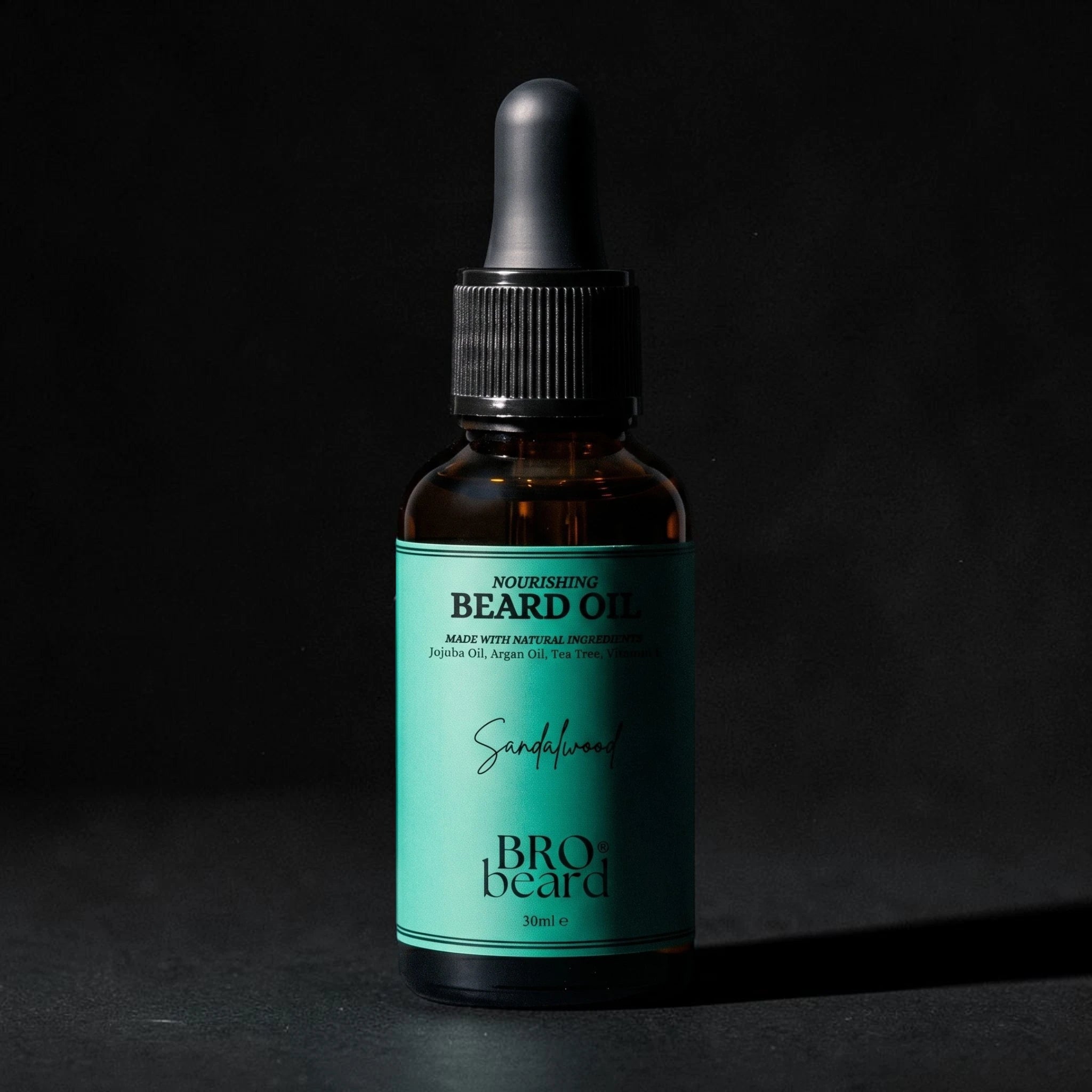 A small amber glass dropper bottle of beard oil with a black cap and a bright teal label reading 'Bro beard' and 'Beard Oil', photographed against a dark black background.